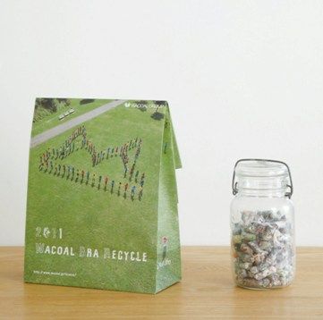 A green + grass-print paper bag with "Wacoal Bra Recycle" printed on it sits on a wooden surface. A clear jar of what looks like recovered fibre sits to the right of the bag.